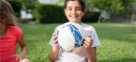 En glad flicka i vit tröja sitter på marken. I famnen håller hon en vit och blå boll. 