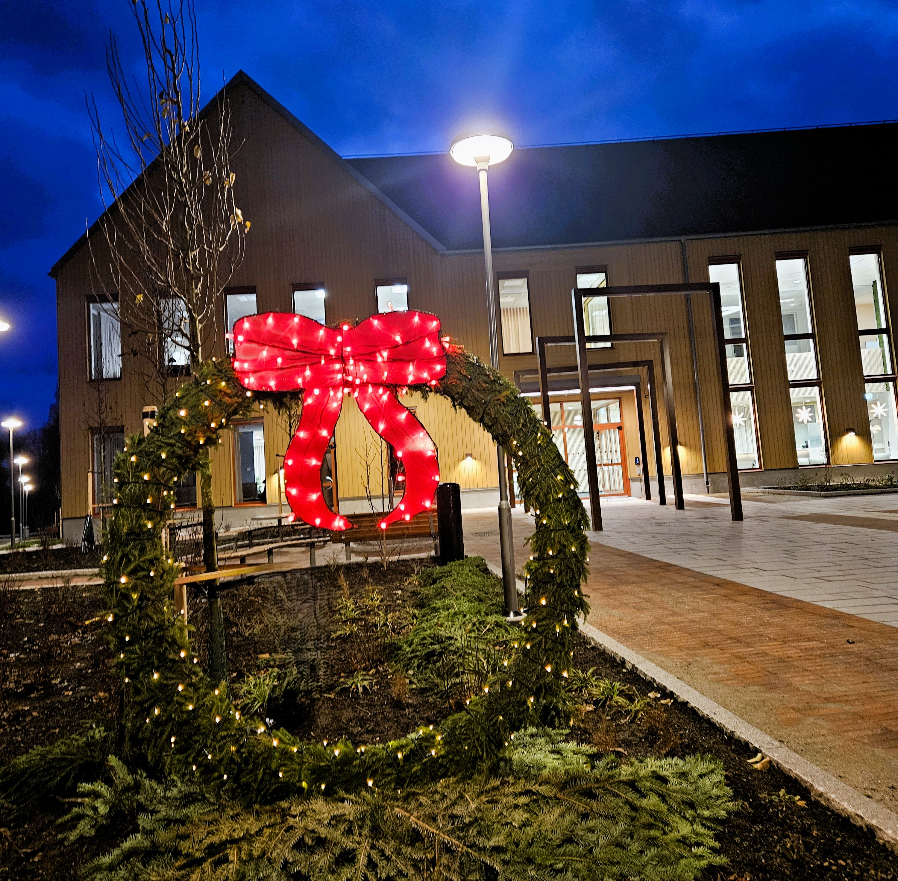 En stor julkrans av granris med röd rosett . I bakgrunden finns entren till  kommunhuset i Broby