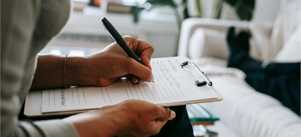 en hand som håller en penna som skriver på ett papper. Bakgrunden är suddig